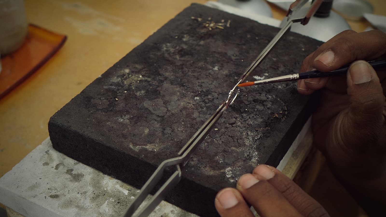 A jeweler's hands brush flux onto a small silver join held in tweezers on a charred soldering block.