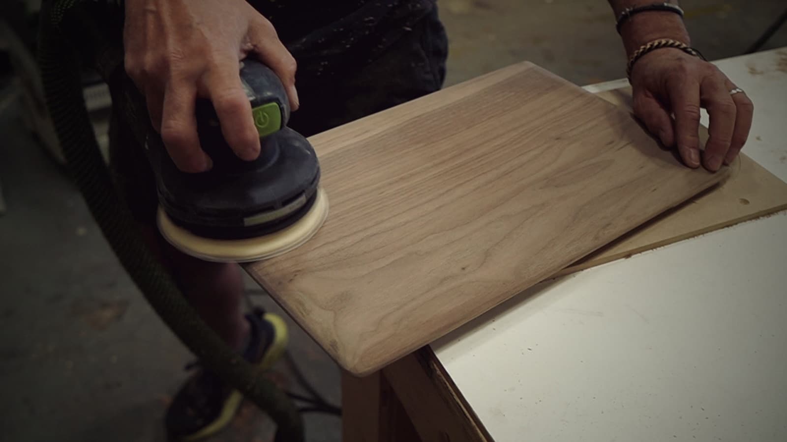 A maker's hands guide an orbital sander across a walnut board at the bench.