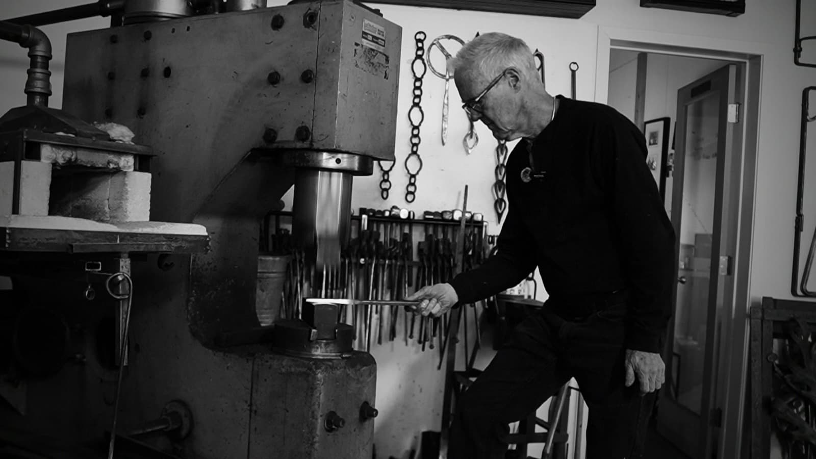 A blacksmith guides a length of hot steel beneath the die of a power hammer, in front of a wall of hanging forging tongs.
