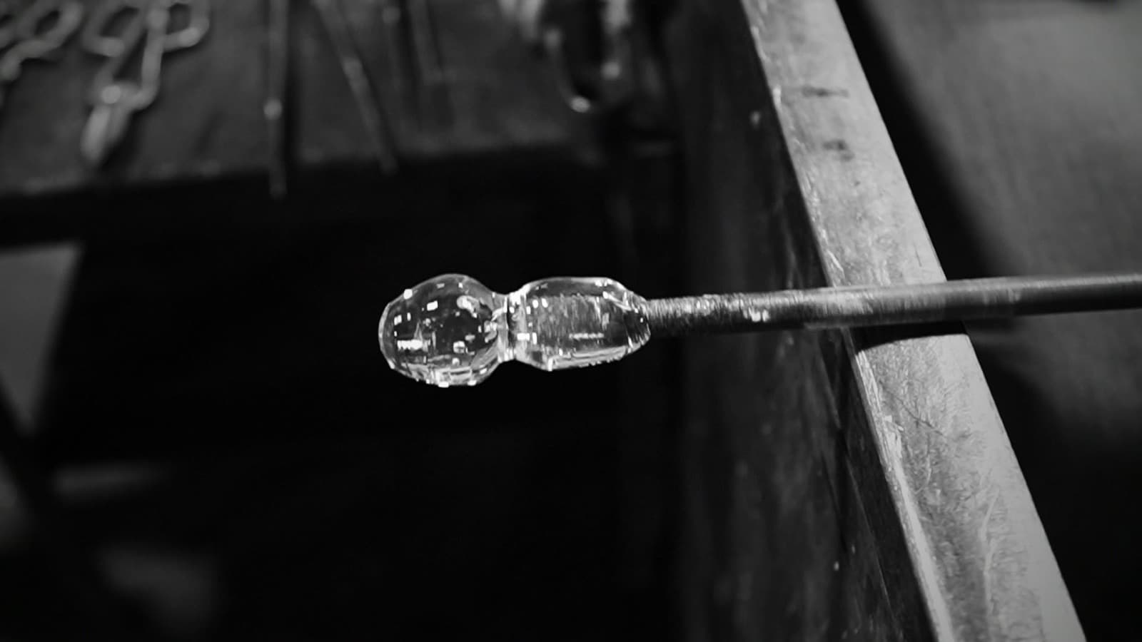 A cooling gather of glass rests on the end of a blowpipe above the bench, jacks and shears laid out behind it.