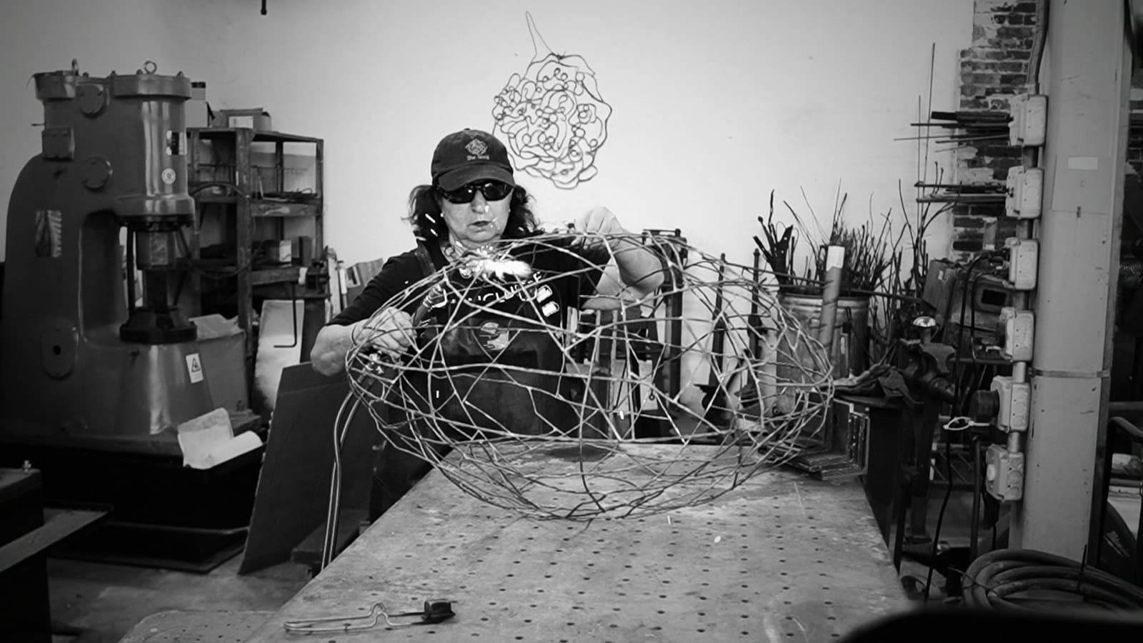 A blacksmith in a cap and safety glasses torches a joint on a large forged-wire globe sculpture at her steel worktable, a power hammer and racks of stock behind her.