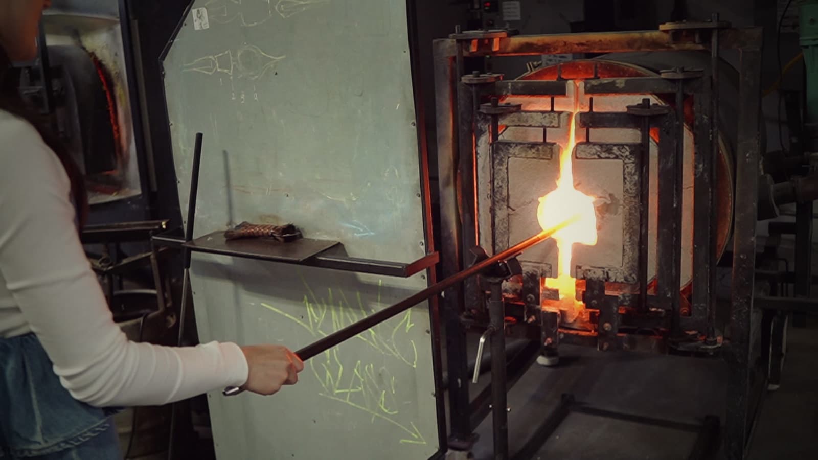 A glassblower reaches a blowpipe into the mouth of a glory hole, the gather at its tip glowing white-hot.