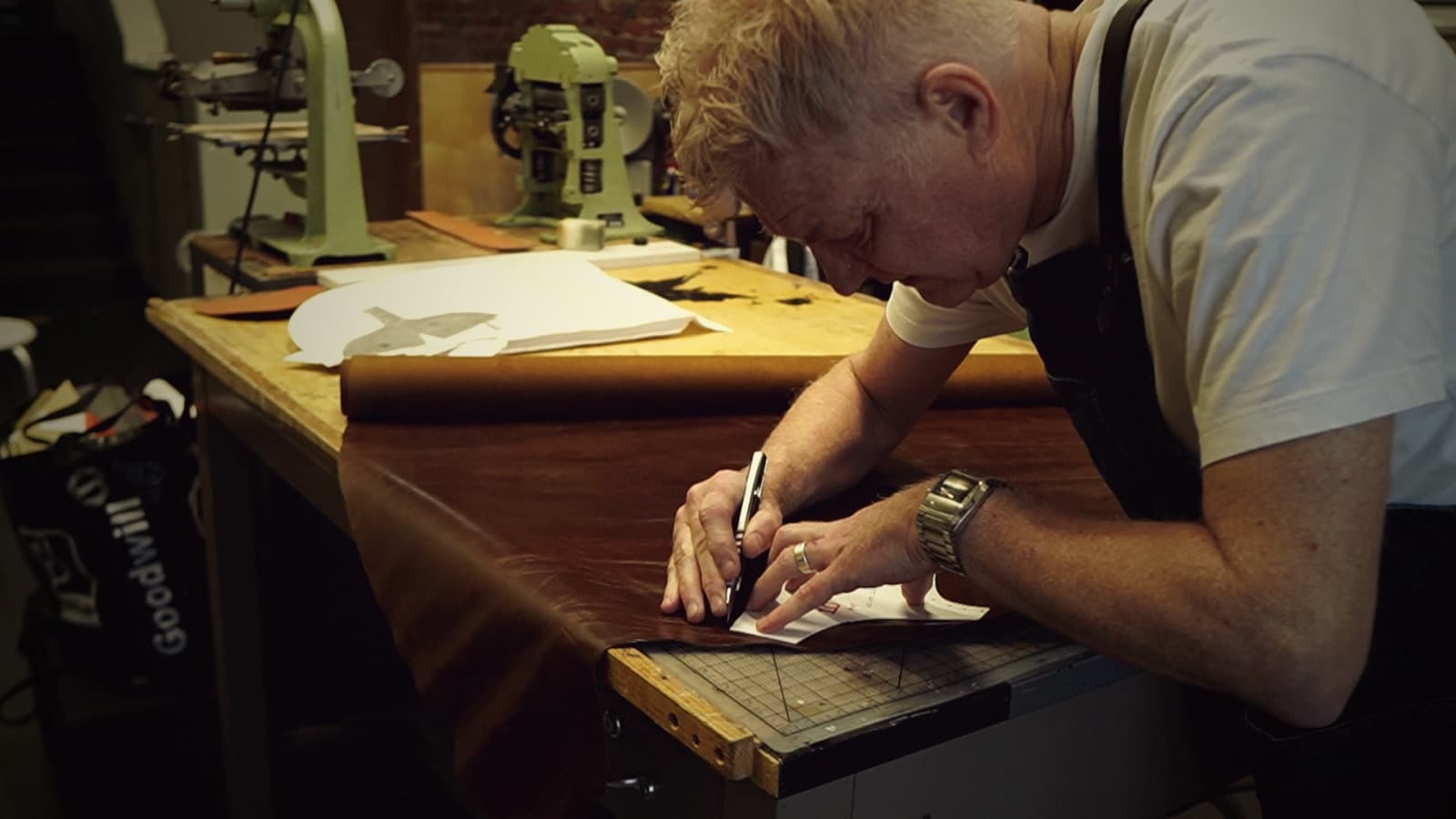 A shoemaker leans over the bench, cutting a pattern piece from a hide of brown leather with a knife.