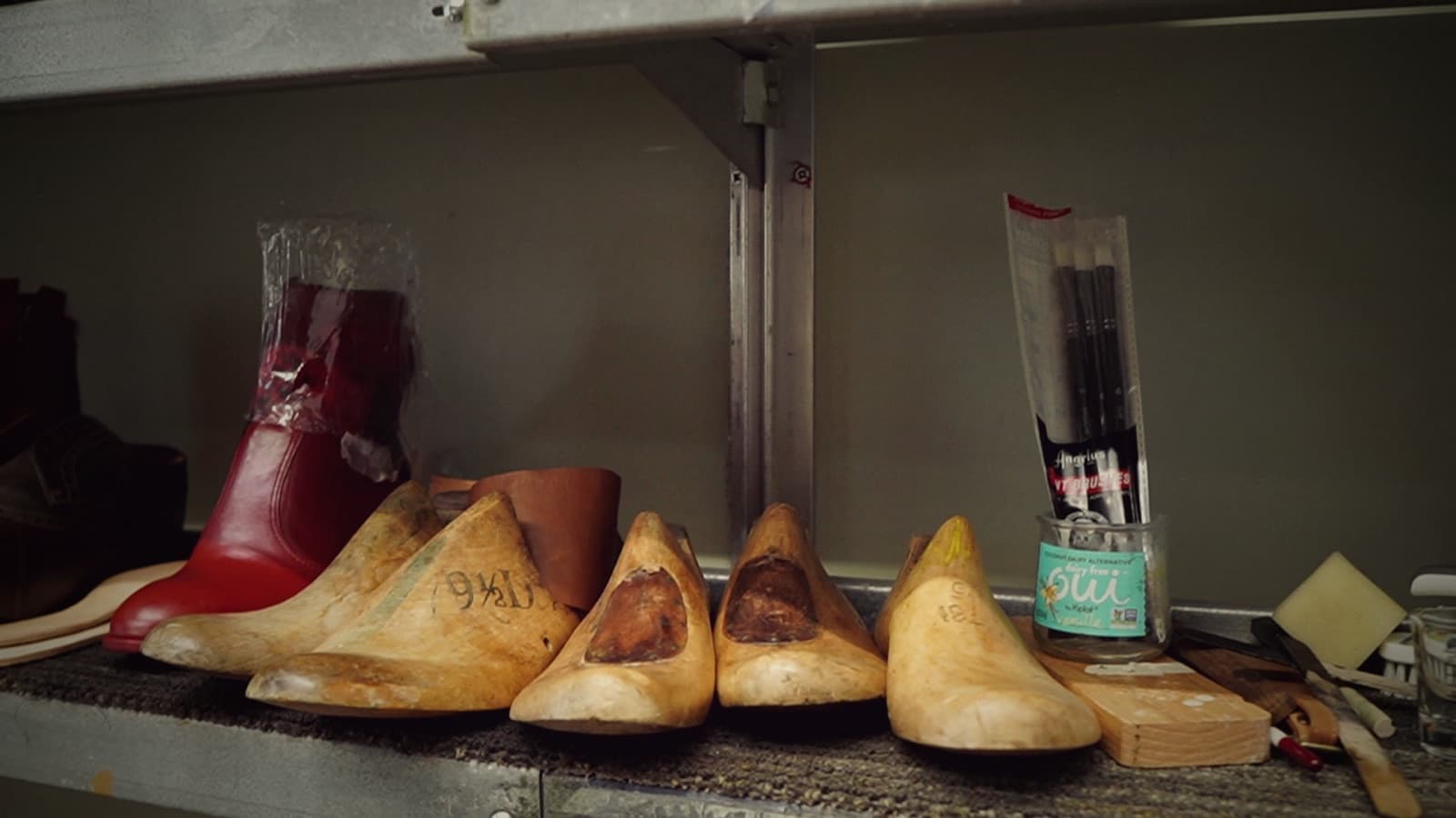 Wooden shoe lasts and a red leather boot line a cobbler's shelf beside brushes and hand tools.