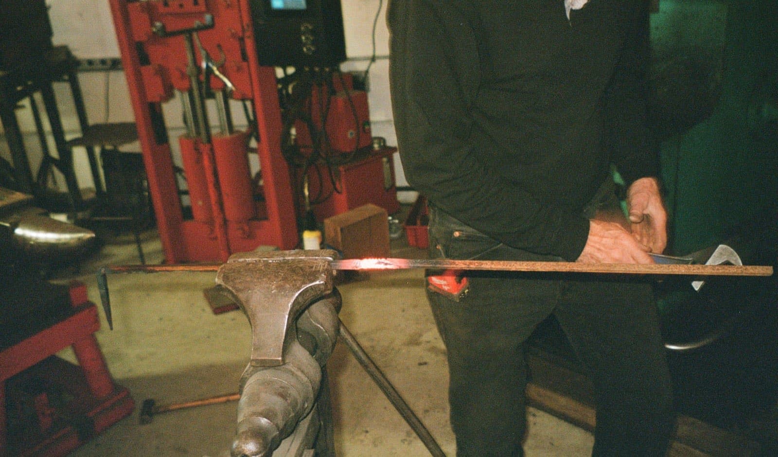 A blacksmith working a long steel bar, glowing red where it crosses the anvil